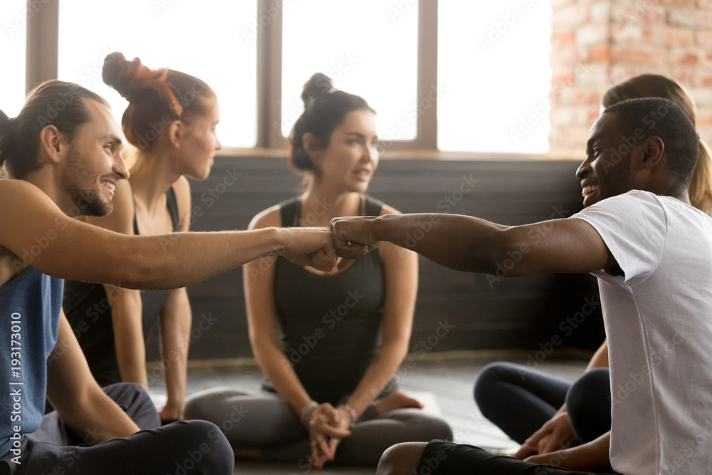Smiling sporty fit african american and caucasian men fist bumping at group training meeting in gym studio, two diverse friends greeting or supporting team motivation for sport yoga teamwork concept Smiling sporty fit african american and caucasian men fist bumping at group training meeting in gym studio, two diverse friends greeting or supporting team motivation for sport yoga teamwork concept