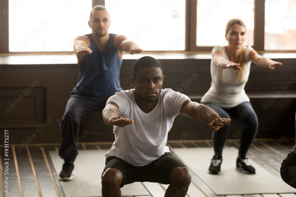 Young african american man doing squat exercise at group fitness training,  sporty black guy focused on self-improvement working out with diverse  active people in gym studio during routine session Stock Photo