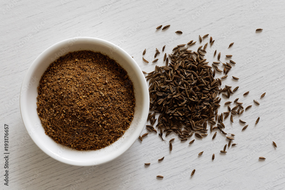Ground cumin in white ceramic bowl isolated on white wood background from above. Whole cumin. Ground cumin in white ceramic bowl isolated on white wood background from above. Whole cumin.