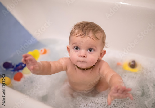 Curious Baby boy is looking over the camera while taking the bath with foam. Drawing his hands to the mother 