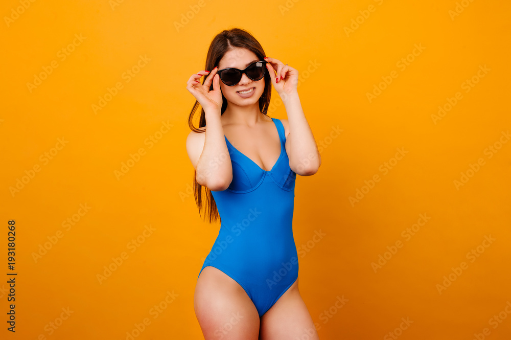 Portrait of happy smiling young beautiful woman in body blue swimsuit touching sunglasses. Slim sexy young woman in swimsuit over orange background.