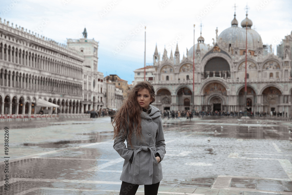 Beautiful tourist girl with long wavy hair in San Marco square in front of the basilica, Venice, Italy. Happy smiling brunette wearing in gray coat enjoying holiday.