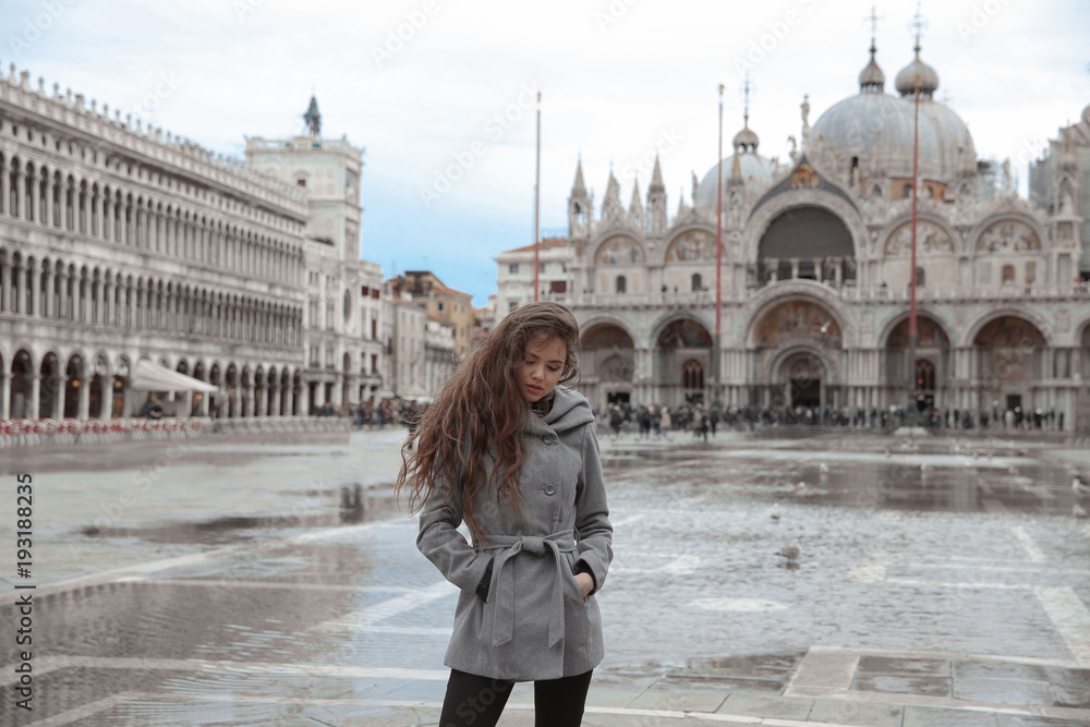 Beautiful tourist girl with long wavy hair in San Marco square in front of Saint Mark's Basilica, Venice, Italy. Smiling brunette traveller wearing in gray coat enjoying holiday.