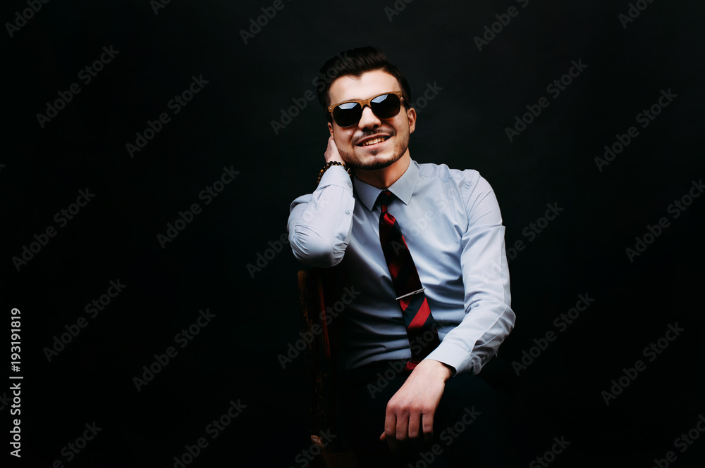 
Handsome smiling and cheerful young business man in casual sitting on chair and wearing sunglasses and looking at the camera over dark background. - obrazy, fototapety, plakaty 