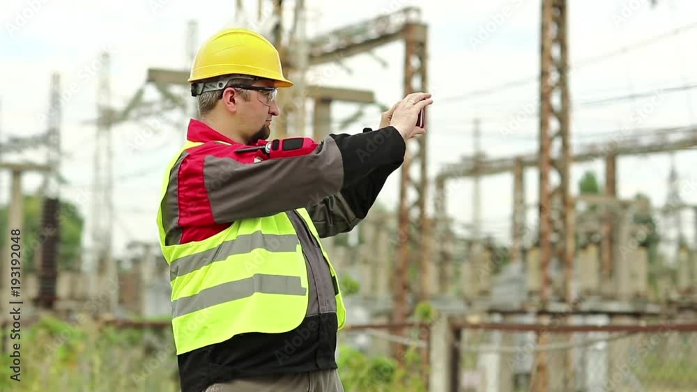 Electrician records videos on smartphone. Powerman in yellow hard hat with smartphone at electric power plant Electrician records videos on smartphone. Powerman in yellow hard hat with smartphone at electric power plant