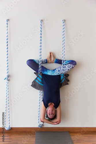 Iyengar yoga practitioner hanging upside down from ropes on white wall. Yogi doing calisthenics exercise to stretch body. Difficult physical pose, bat posture concept