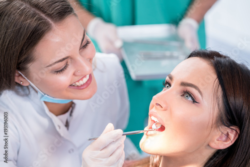 people, medicine, stomatology and health care concept - happy female dentist with mirror checking patient girl teeth up at dental clinic office while male assistant is waiting behind.