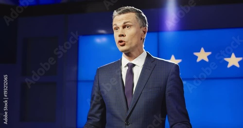 Mature politician in elegant suit standing on stage in light against screen with EU flag and giving speech while arranging press conference.