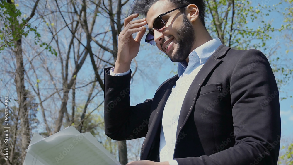Muslim man picks up papers in hands and smiles. Male person stands in park and dressed Muslim man picks up papers in hands and smiles. Male person stands in park and dressed