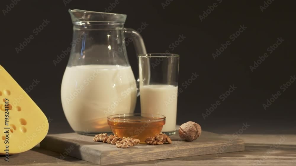 Wedge of Swiss cheese with jug of milk on an old wooden table. Close-up of board with cheese, milk jug, nuts and honey on a dark background. Dolly shot Wedge of Swiss cheese with jug of milk on an old wooden table. Close-up of board with cheese, milk jug, nuts and honey on a dark background. Dolly shot