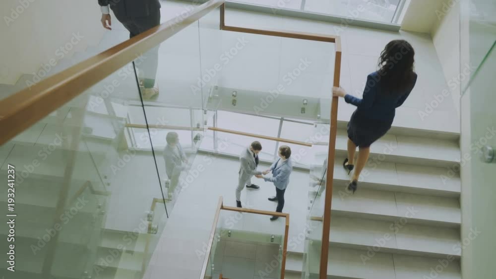Top view of two businessmen meet at staircase in modern office center and talking while female colleagues walking stairs Top view of two businessmen meet at staircase in modern office center and talking while female colleagues walking stairs