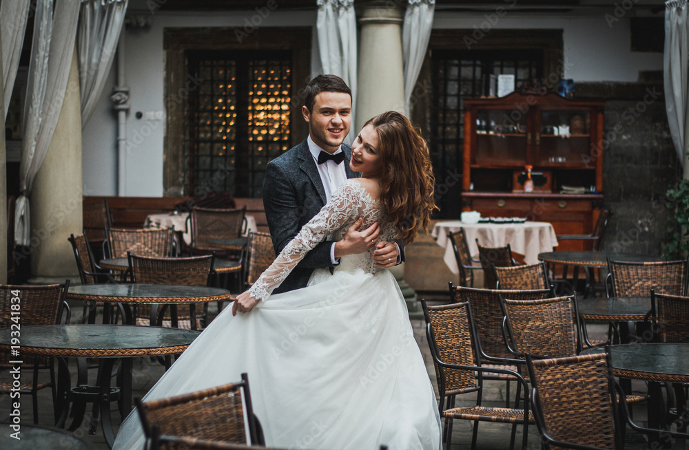 Wedding couple is walking in the old city. Stone walls of ancient town on background. Bride with streaming hair and groom in grey suit and bow tie. Lace elegant dress. Romantic love. Smile emotions. Wedding couple is walking in the old city. Stone walls of ancient town on background. Bride with streaming hair and groom in grey suit and bow tie. Lace elegant dress. Romantic love. Smile emotions.