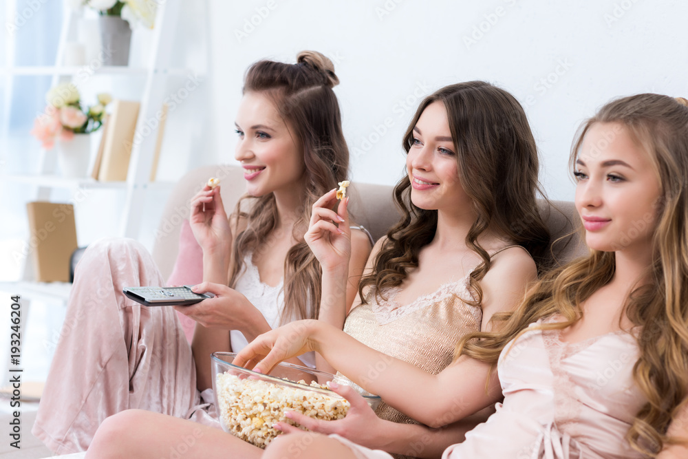 beautiful smiling girlfriends in pajamas eating popcorn and watching tv together