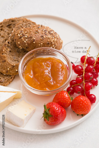 Top view on white plate with slices of organic wholegrain bread, Camembert cheese, apricot jam, red current and strawberries. Healthy snack or breakfast at home.