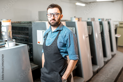 Portrait of a handsome typographer standing at the printing manufacturing with offset machine on the background