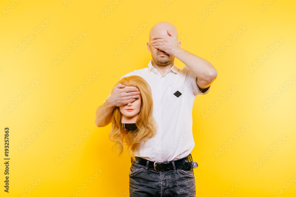 Excited odd bizarre strange unusual sad adult bald hairless man holding plastic blonde woman head. Funny male with girlfriend isolated on yellow. Baldness after chemotherapy. Cancer hair loss concept. Excited odd bizarre strange unusual sad adult bald hairless man holding plastic blonde woman head. Funny male with girlfriend isolated on yellow. Baldness after chemotherapy. Cancer hair loss concept.