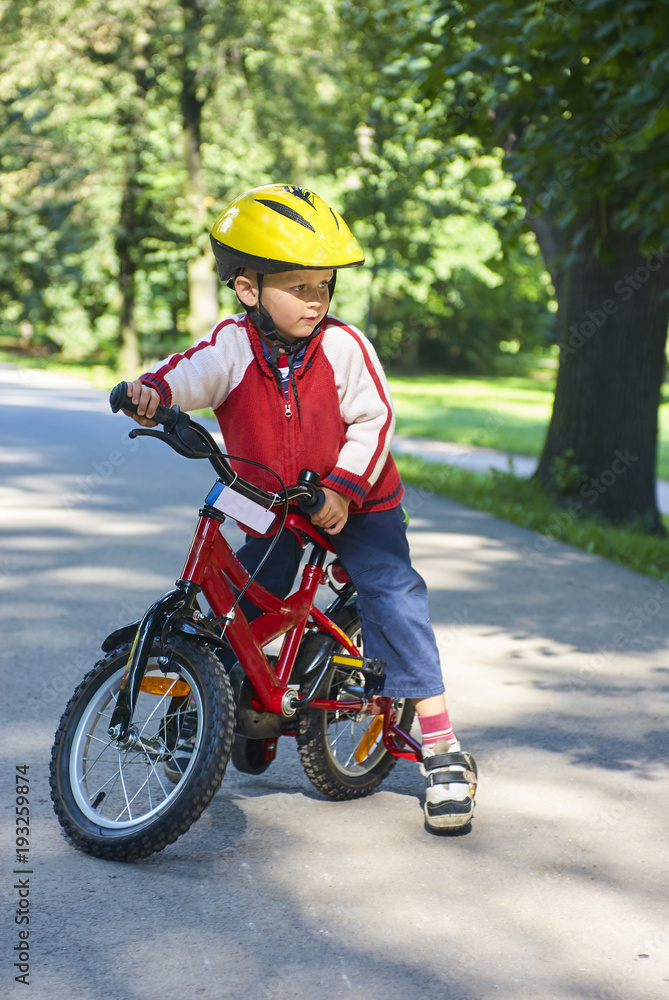 Little boy on bicycle in green park outdoor in summer. A child is riding a children's bike with support wheels wearing safety helmet Little boy on bicycle in green park outdoor in summer. A child is riding a children's bike with support wheels wearing safety helmet