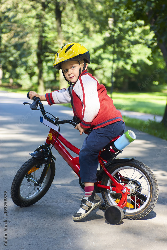 Little boy on bicycle in green park outdoor in summer. A child is riding a children's bike with support wheels wearing safety helmet Little boy on bicycle in green park outdoor in summer. A child is riding a children's bike with support wheels wearing safety helmet
