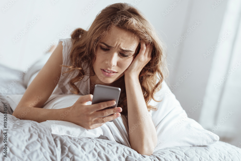 Trying to find an answer. Depressed mature lady resting her head on a hand and grimacing while lying on a bed and looking at her phone. Trying to find an answer. Depressed mature lady resting her head on a hand and grimacing while lying on a bed and looking at her phone.