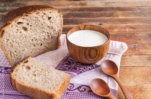A wooden cup of milk and bread on lilac kitchen towel on old wooden table.