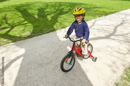 Little boy on bicycle in green park outdoor in summer. A child is riding a children's bike with support wheels wearing safety helmet
