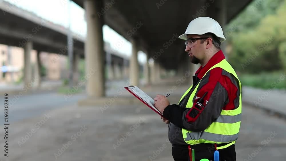 Road mender makes notes in work documentation. Building inspector with work documentation at project site under overpass Road mender makes notes in work documentation. Building inspector with work documentation at project site under overpass