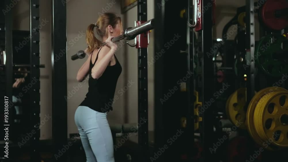 young girl with white hair in black tank top and blue tights doing squats with a vulture from a barbell in a dark fitness club young girl with white hair in black tank top and blue tights doing squats with a vulture from a barbell in a dark fitness club