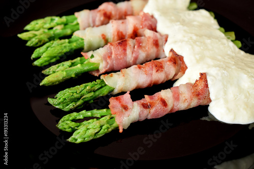 Green asparagus wrapped into smoked bacon covered with white sauce on a black plate