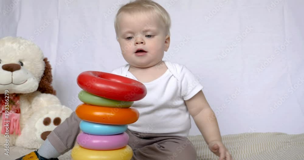 Portrait of a beautiful little kid (boy), sits playing toy pyramid, blond, light-haired, blue eyes, white background. Concept: babies, fair skin, beautiful children. Portrait of a beautiful little kid (boy), sits playing toy pyramid, blond, light-haired, blue eyes, white background. Concept: babies, fair skin, beautiful children.