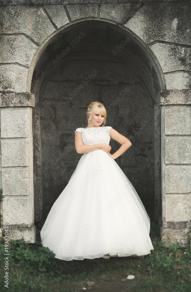 Obraz premium Beautiful elegant bride with perfect wedding dress and bouquet posing near old castle