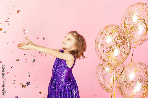 Happy young girl in purple glittering dress celebrating with hands up and smile or skreaming with joy while cathcing confetti, having fun on pink background. photo