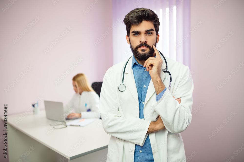 Portrait of young serious male doctor standing in front of camera, male doctor in background working on the computer. Portrait of young serious male doctor standing in front of camera, male doctor in background working on the computer.