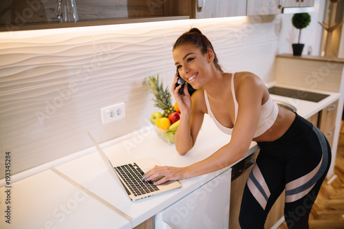 Fit sporty girl using laptop on kitchen table and talking on mobile.