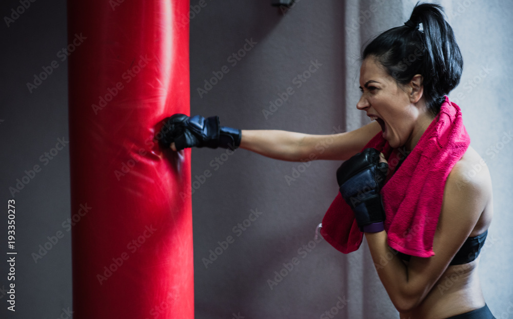 Side view of strong athlete brunette female with red towel on neck punching the red bag in kickboxing gloves at the gym. Woman boxer workout. Sport, fitness, lifestyle, people and motivation concept.