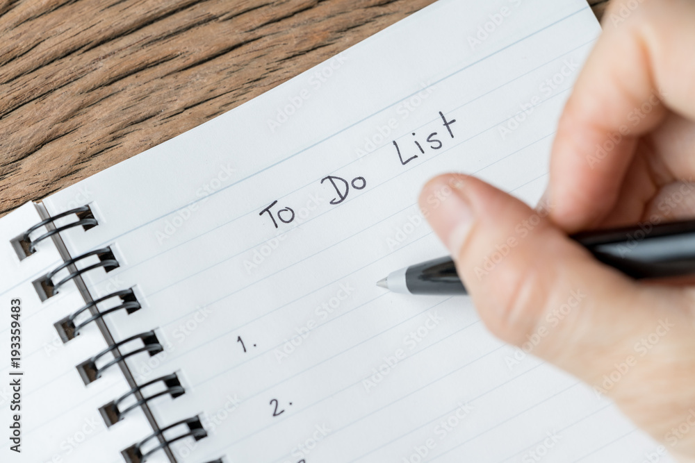 Female hand holding black pen writing To Do List prioritized by number on white paper notepad on wood table in soft tone