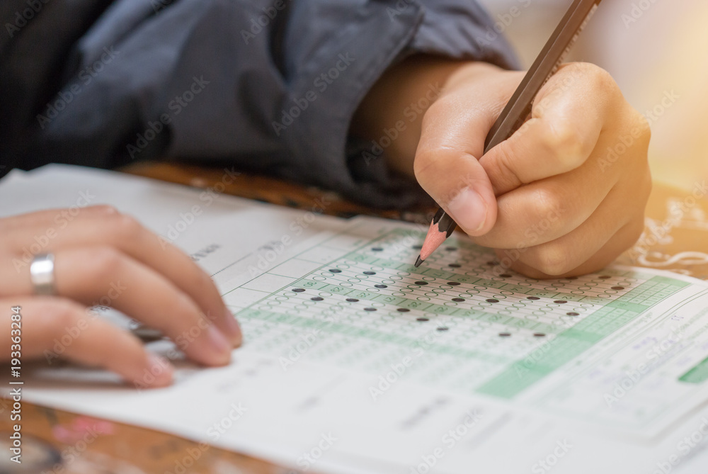 high school or university student hands taking exams, writing examination on paper answer sheet optical form of standardized test on desk doing final exam in classroom. Education literacy concept. high school or university student hands taking exams, writing examination on paper answer sheet optical form of standardized test on desk doing final exam in classroom. Education literacy concept.