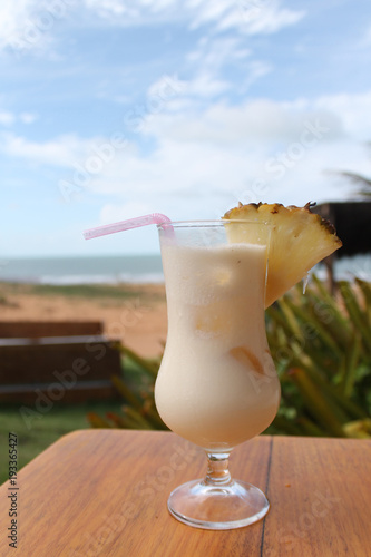 Copo de Piña Colada em uma mesa de madeira com praia ao fundo. Aproveite a vibe tropical!