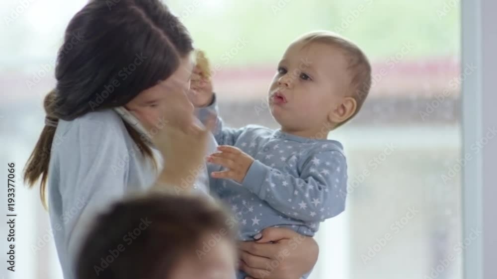 Mother holding baby and talking on the phone and smiling while another child shows up and looks at table Mother holding baby and talking on the phone and smiling while another child shows up and looks at table