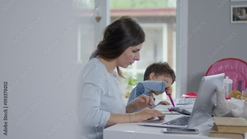 Mother using laptop and smartphone for work and looking after her son who is sitting next to her and drawing Mother using laptop and smartphone for work and looking after her son who is sitting next to her and drawing