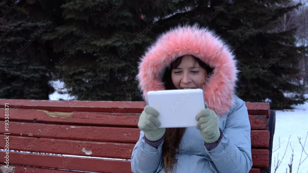 A young woman happy freelancer in a gray warm coat sits on a bench and uses a digital tablet, in a city park in the winter. A young woman happy freelancer in a gray warm coat sits on a bench and uses a digital tablet, in a city park in the winter.