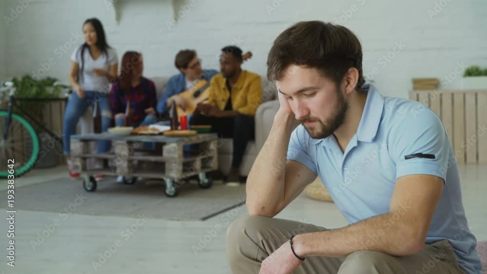 Young student guy feels upset and isolated while his friends celebrating party at home indoors Young student guy feels upset and isolated while his friends celebrating party at home indoors