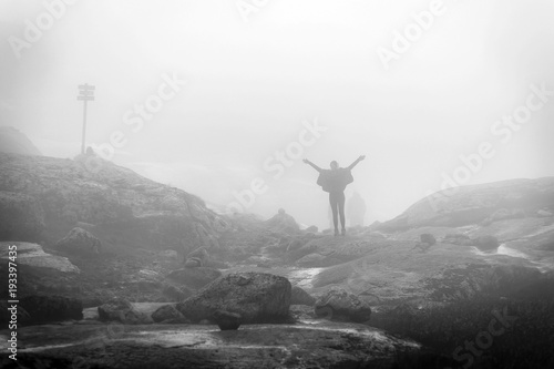 Silhouette of young woman in raincoat walking along the plateau through the foggy place on the way to Kjeragbolten, Norway.