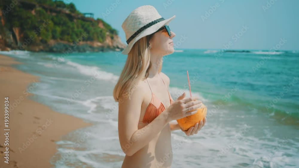 Woman drinks coconut milk and walking on the ocean beach under blue sky in sunny day. Seascape view with palms. Woman drinks coconut milk and walking on the ocean beach under blue sky in sunny day. Seascape view with palms.