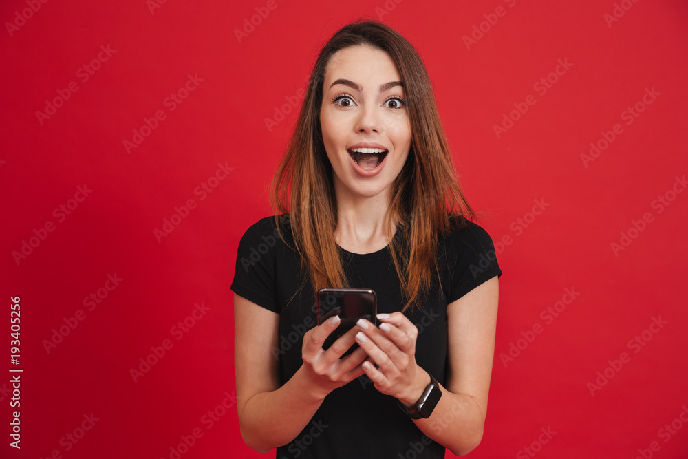 Photo of satisfied woman in black clothing looking on camera while using cell phone with joy, over red background Photo of satisfied woman in black clothing looking on camera while using cell phone with joy, over red background