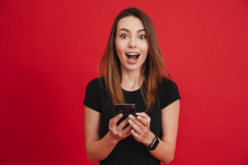 Photo of satisfied woman in black clothing looking on camera while using cell phone with joy, over red background