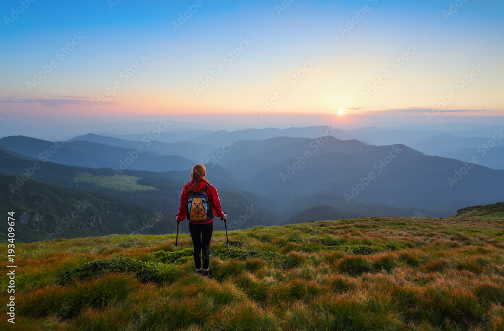 Redheaded girl athlete with a backpack and sticks stands on the green hillocks and looks at high mountain landscapes and fascinating sunset. Location place Carpathian national park, Ukraine, Europe.