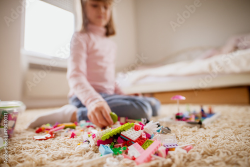 Blurred small toddler girl playing with plastic toy blocks on the carpet in her room.
