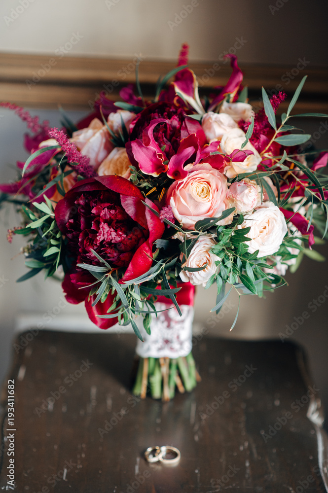 Obraz premium Bridal bouquet in wine red color with lace ribbon on wooden chair, wedding rings near