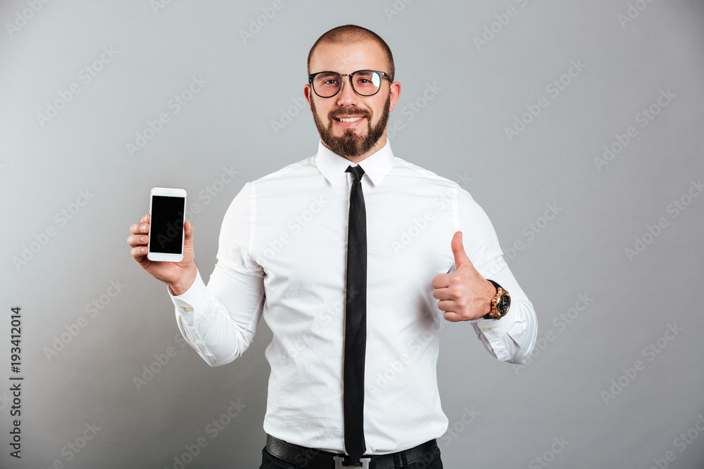 Photo of happy successful man in glasses and tie demonstrating cell phone and showing thumb up, isolated over gray background Photo of happy successful man in glasses and tie demonstrating cell phone and showing thumb up, isolated over gray background