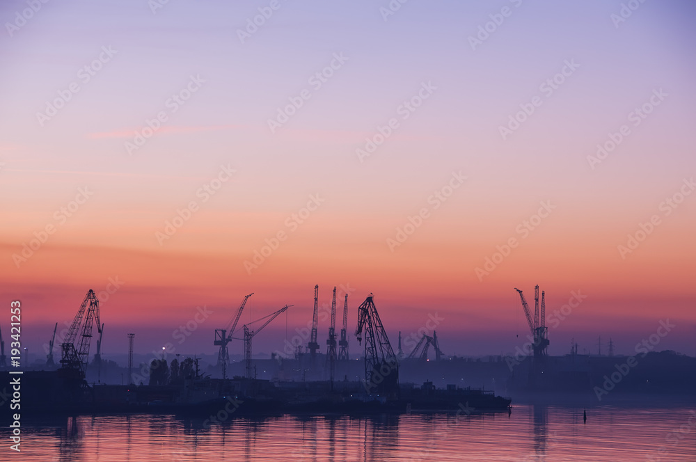 View of the port at sunset.  Industrial warehouse complex on the shore of the baySilhouettes of the cranes in the sunset.
